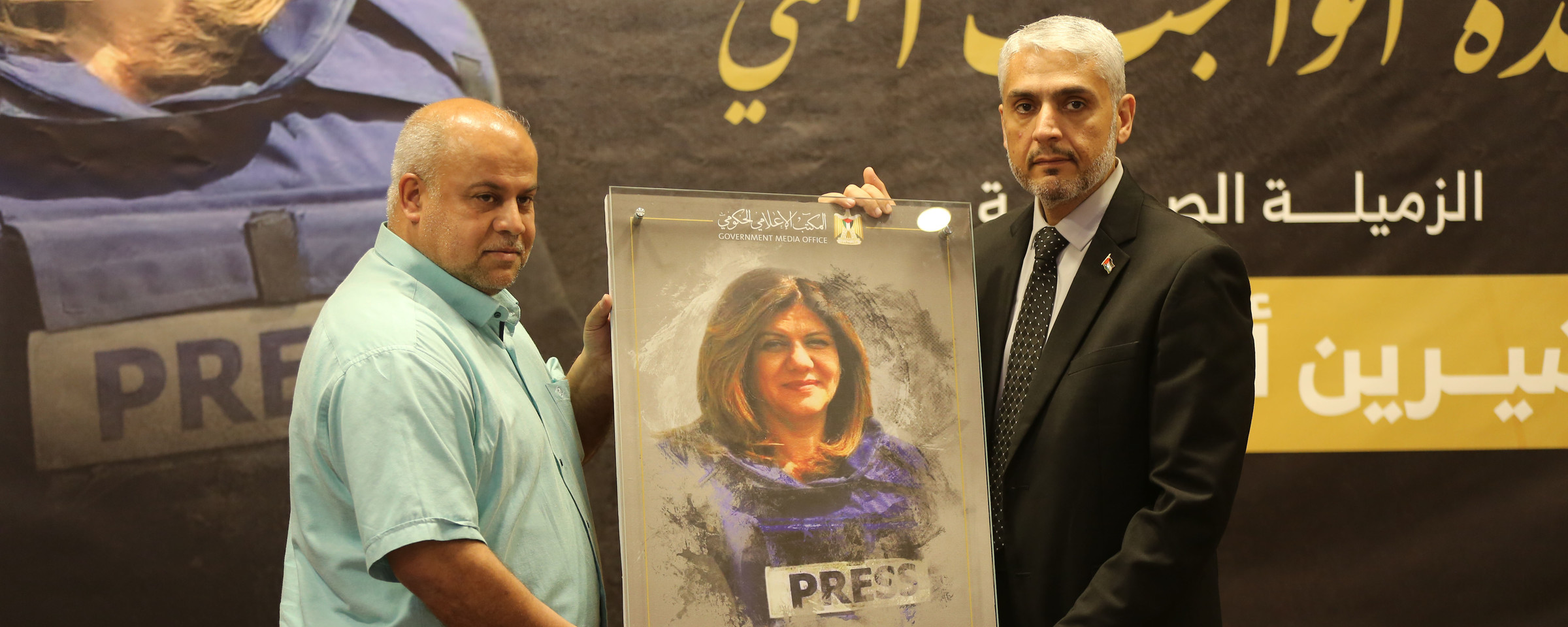 Two men hold framed picture of Shireen Abu Akleh while standing on stage