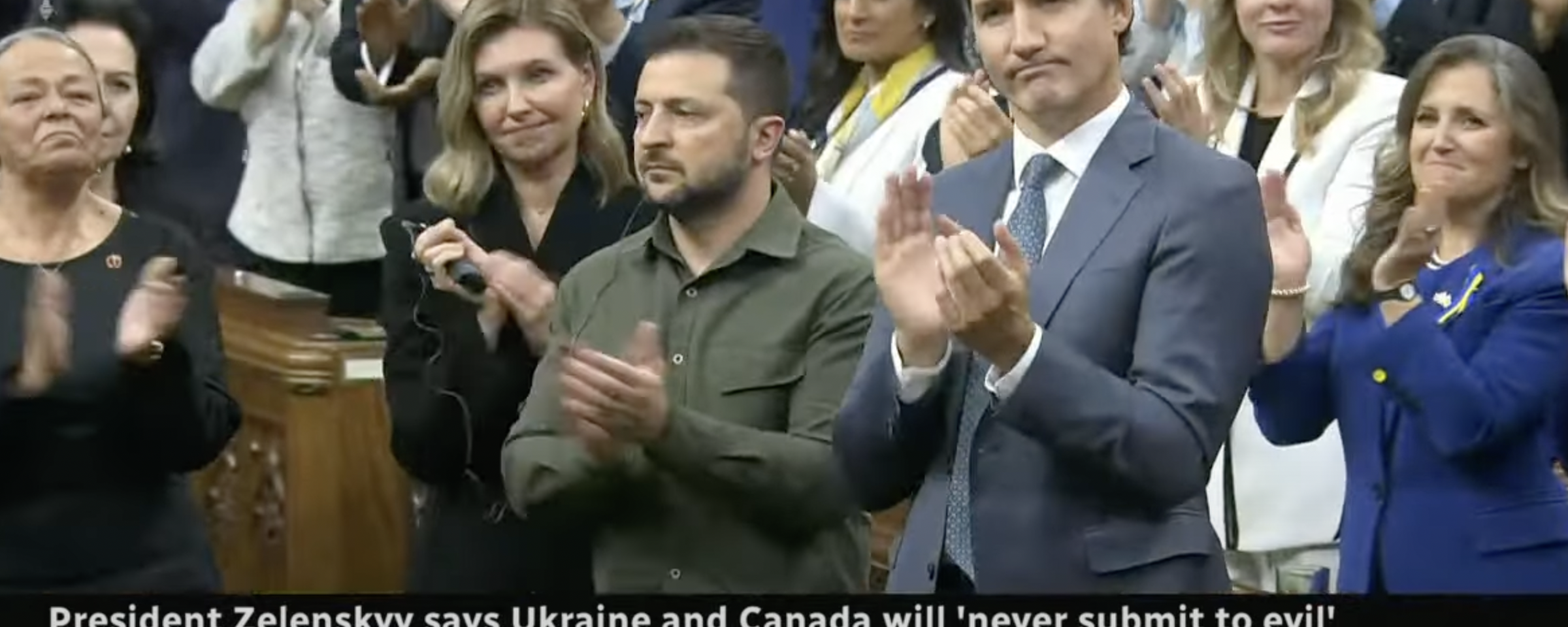 Zelensky in a military shirt stands next to Trudeau who is wearing a suit and clapping as others look on