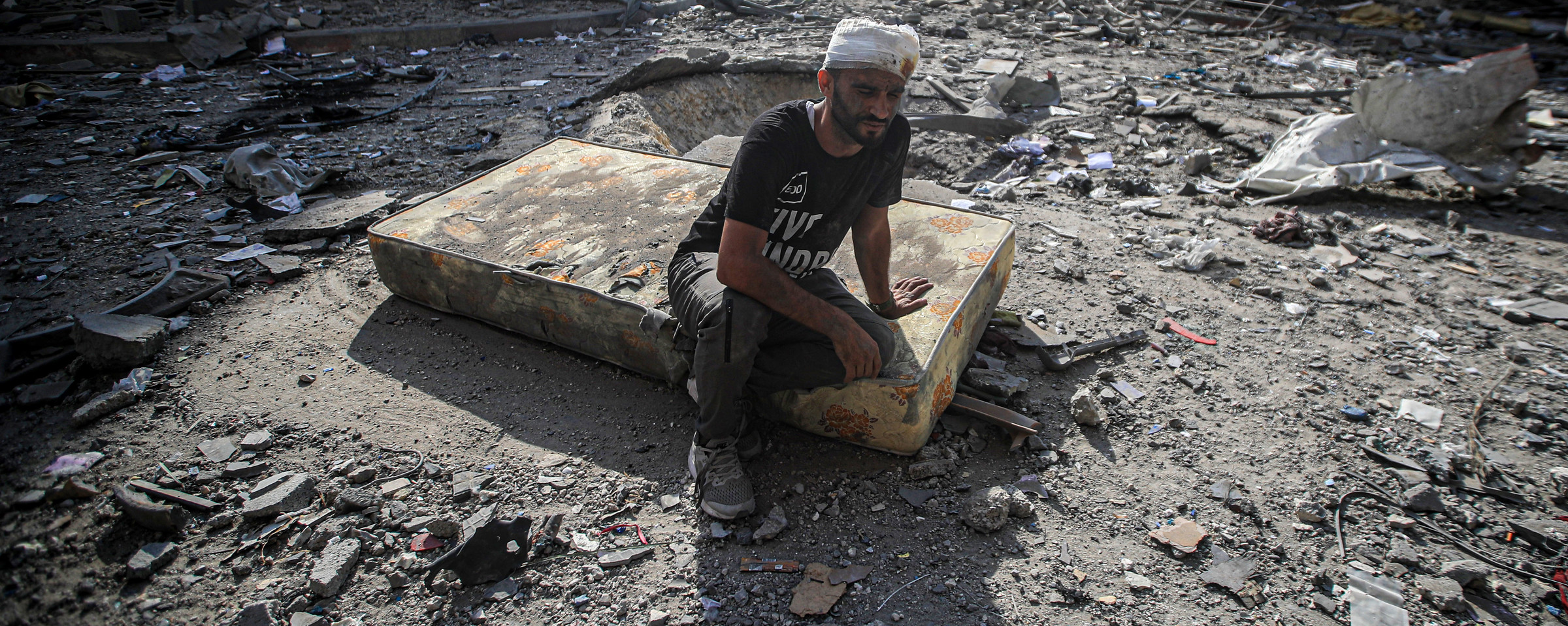 A man with a bandaged head sits on a mattress in a debris-filled street