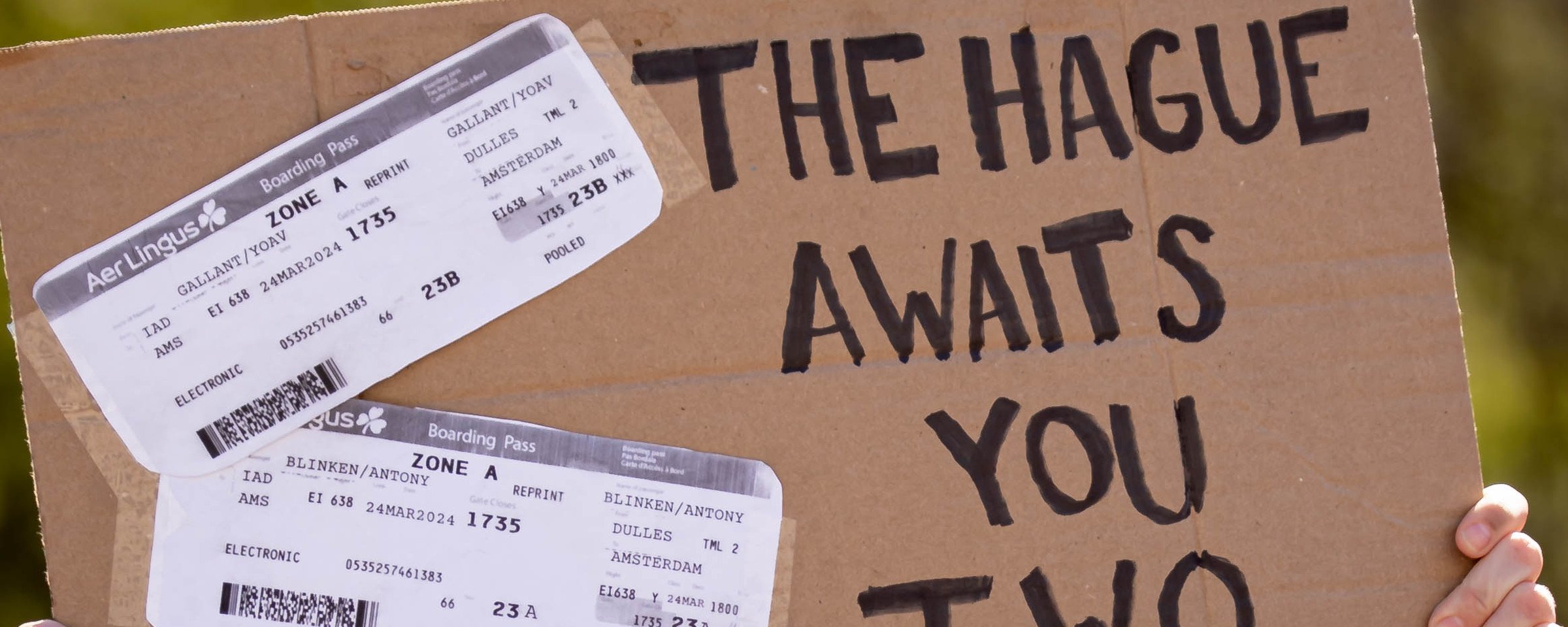 A cardboard protest sign reads "The Hague Awaits You Two" with mock plane tickets for Yoav Gallant and Antony Blinken