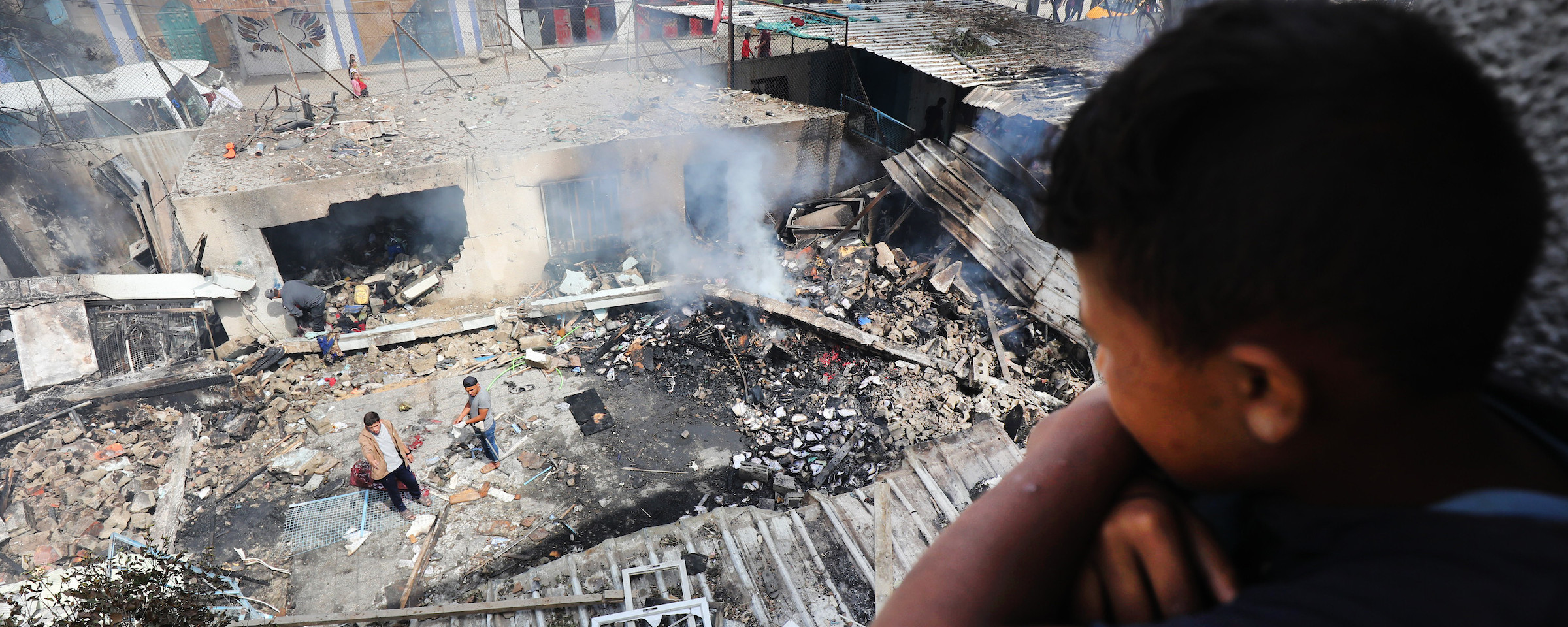 A boy leans over a railing while looking down at smoking debris where a building once stood