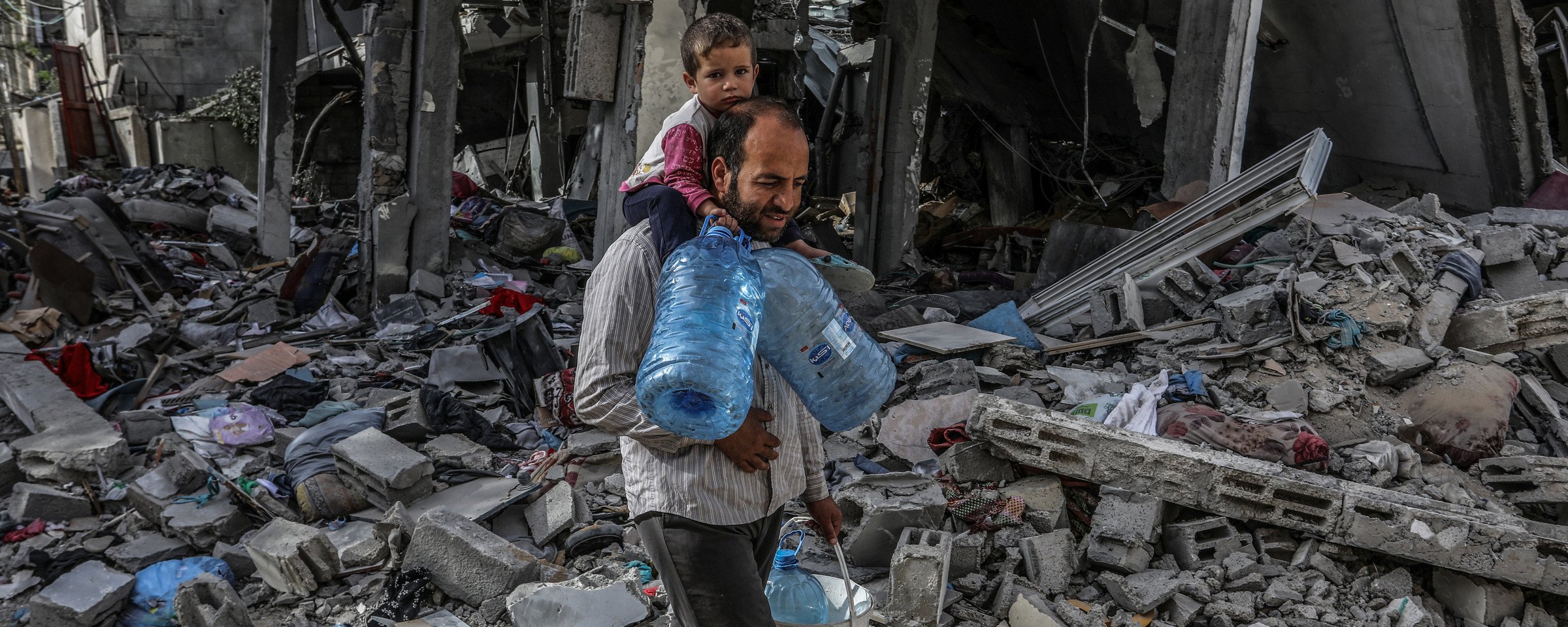 A man carries a child on his shoulders and water containers in his hands as he walks through rubble in southern Gaza 