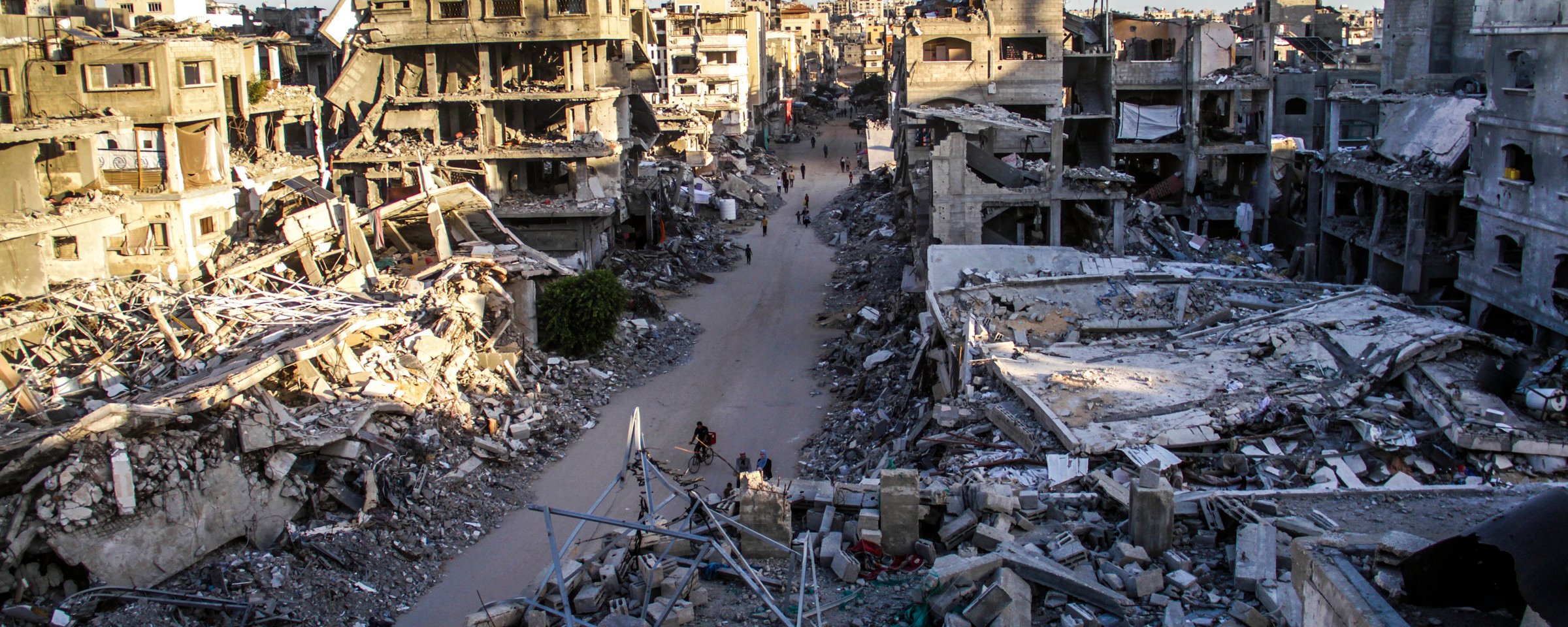 Landscape view of people walking and biking on narrow road cleared of debris between destroyed and bombed-out multi-story buildings