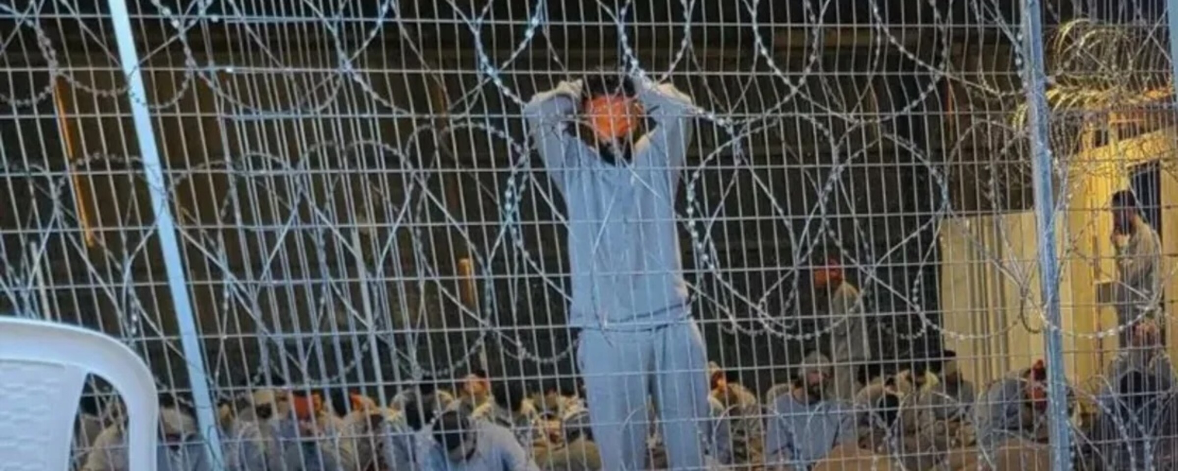 Men blindfolded and bound sit or stand behind razor wire fence