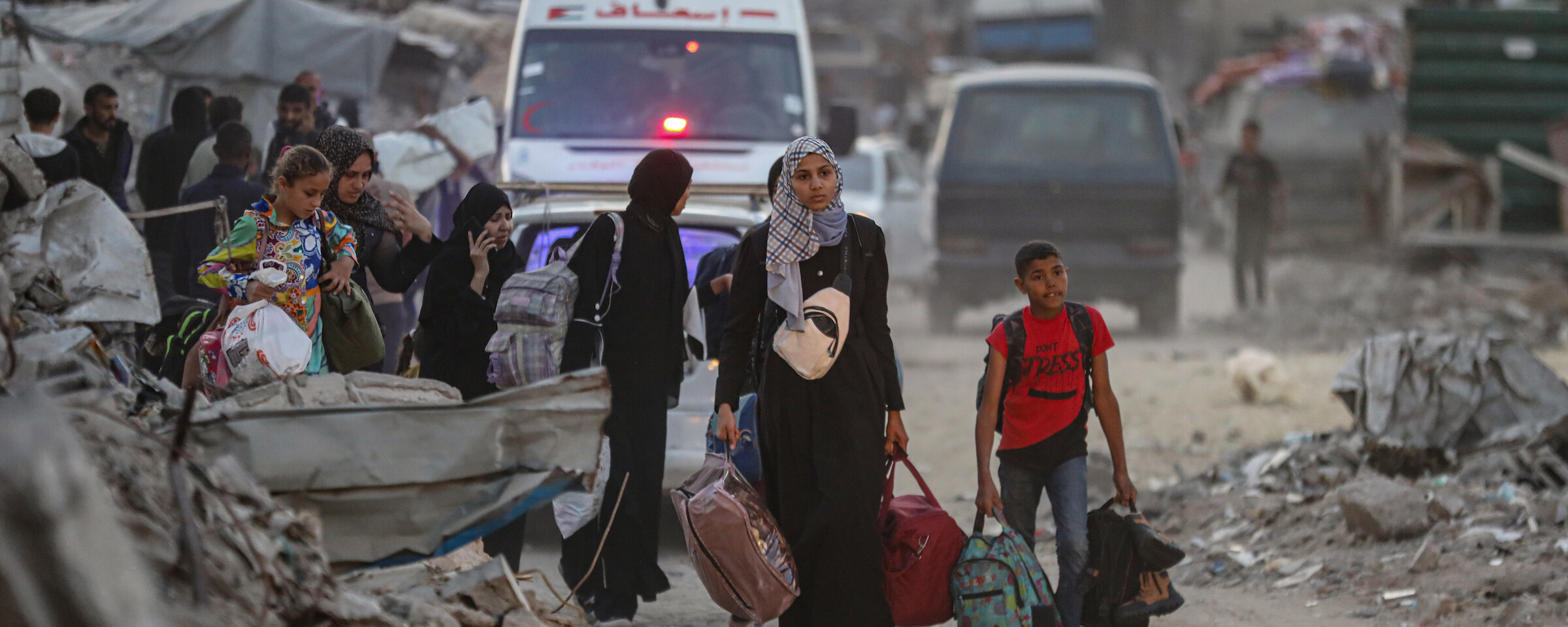 Women and children carry belongings amidst rubble. Ambulances are behind them.