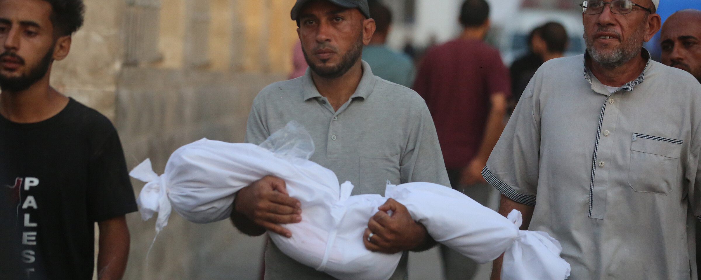 A man holds the body of a child, wrapped in a white burial shroud. Two men walk alongside him