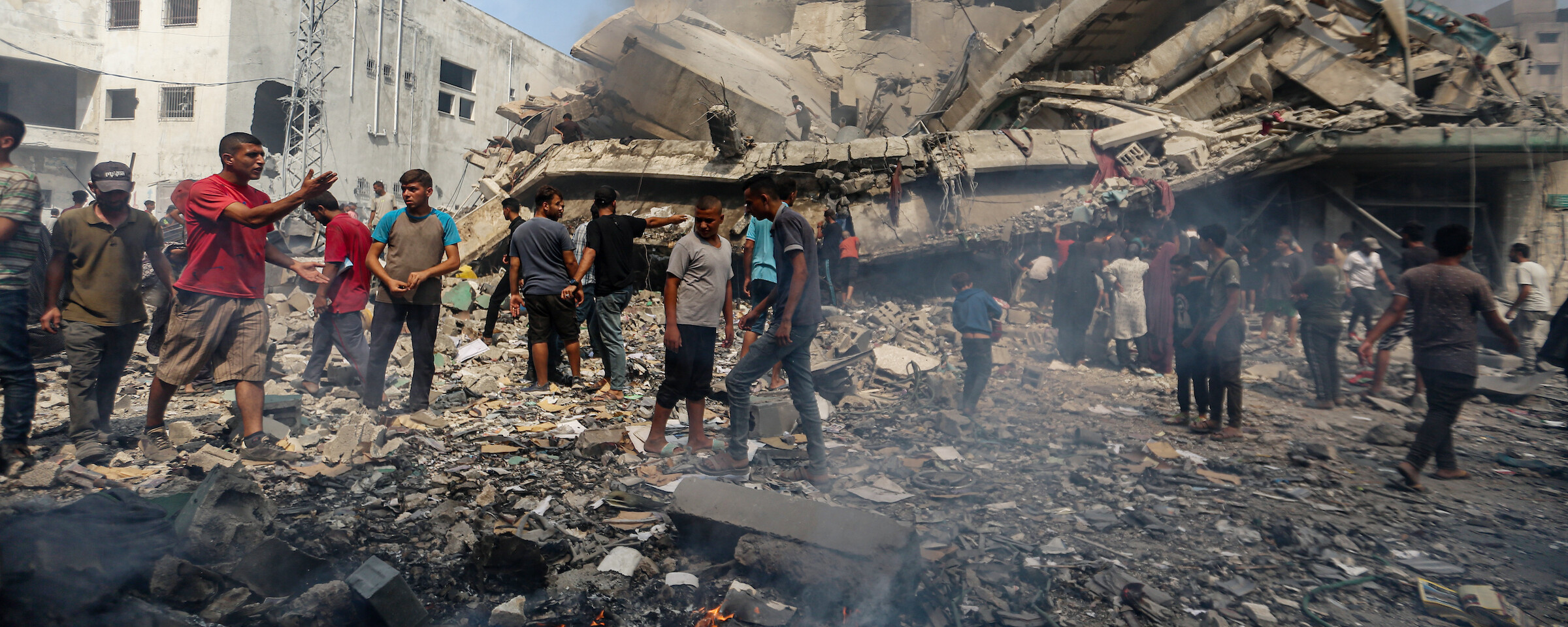 People stand on top of the rubble of a destroyed building, with black smoke rising around them. Several small fires are visible in front of them.