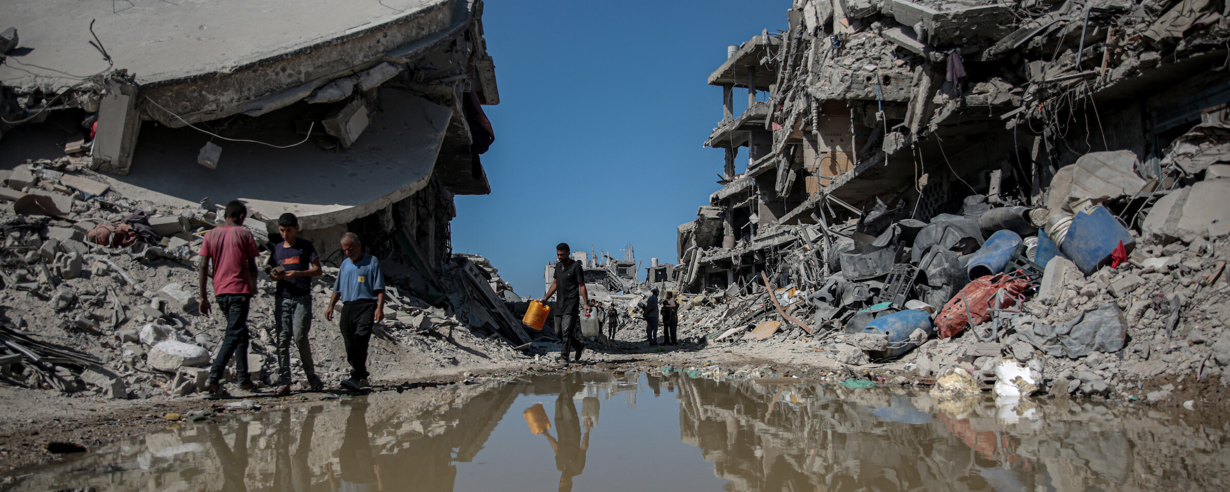 Several Palestinians in Jabaliya walk by standing water and demolished buildings