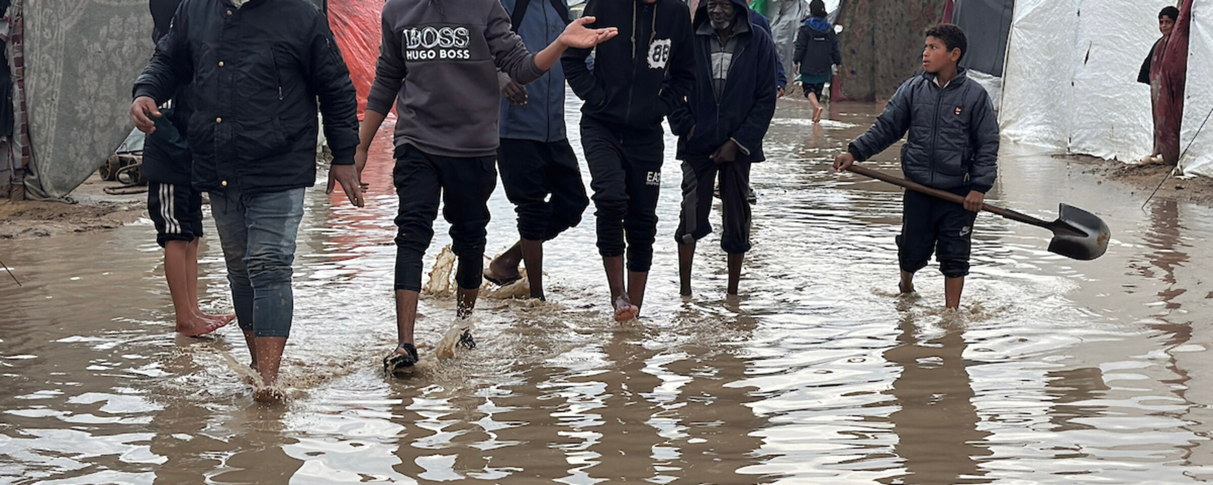 A group of youth walk through a flooded street between tents. One of them is carrying a shovel.