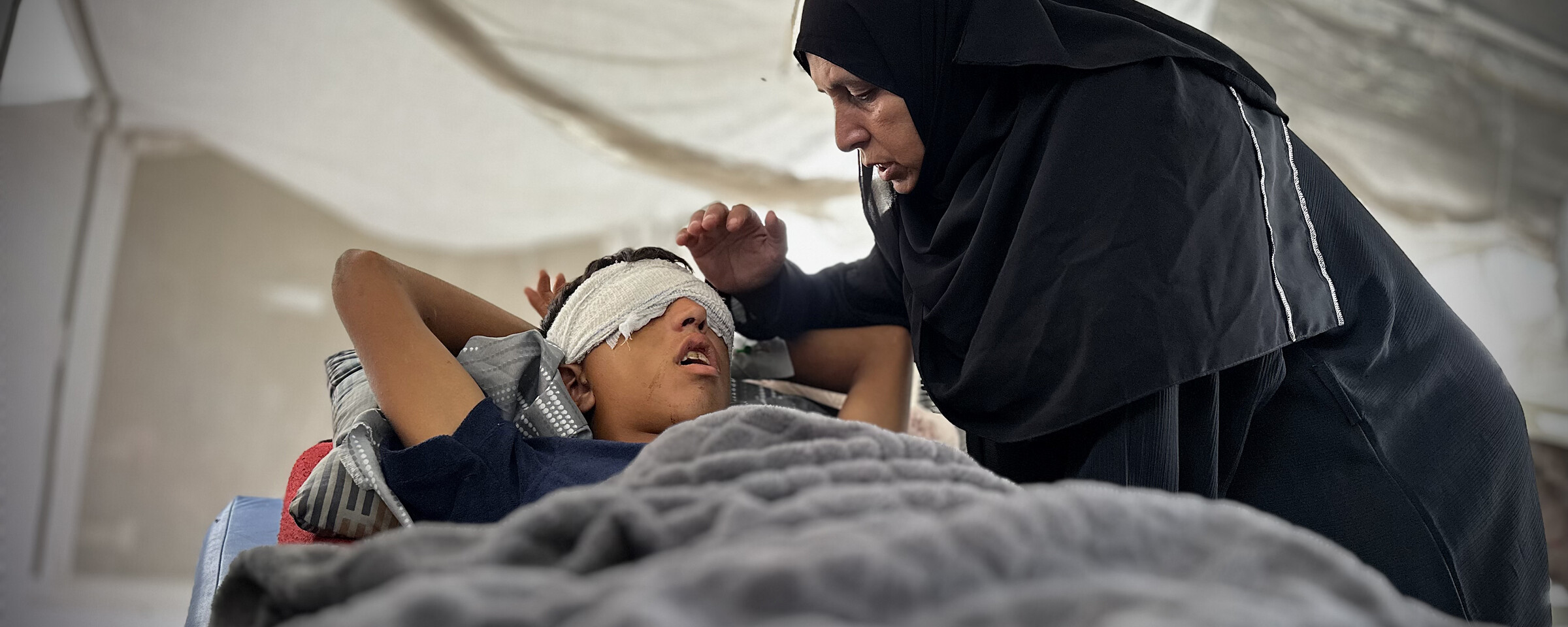 A woman leans and holds her hand over a boy on bed with a bandage wrapped around his eyes and head