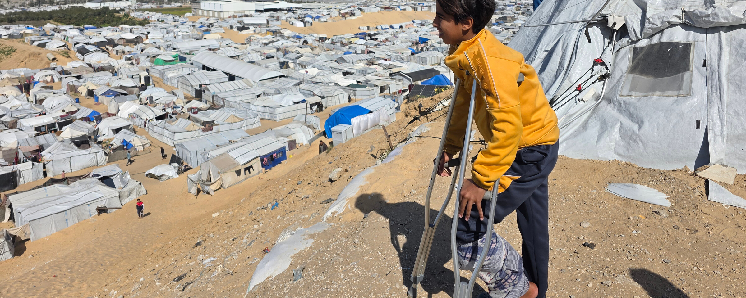 A young boy on crutches stands next to a tent overlooking white tents on sand.