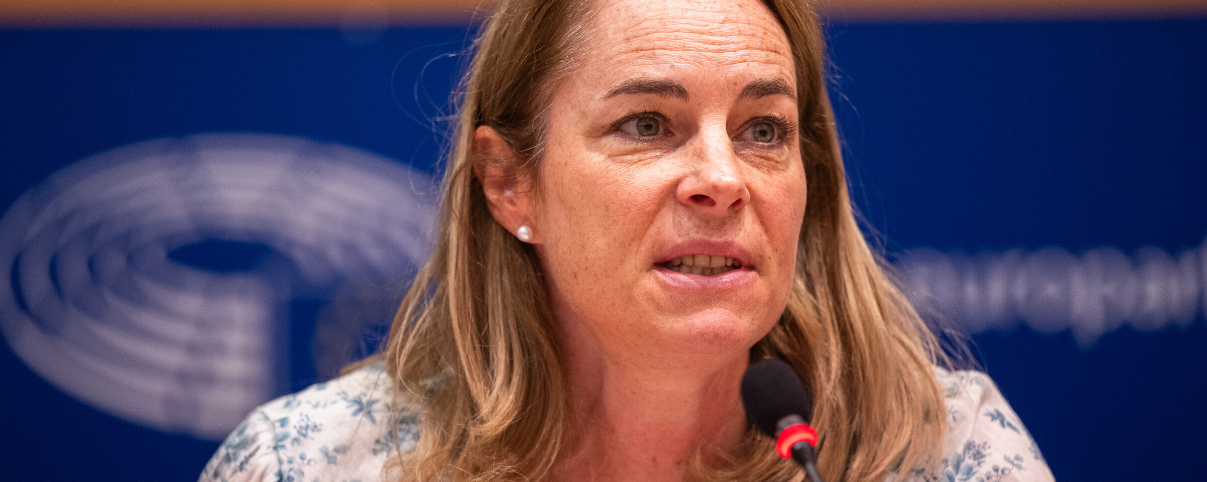 A woman wearing a blouse with a floral pattern speaks into a microphone 