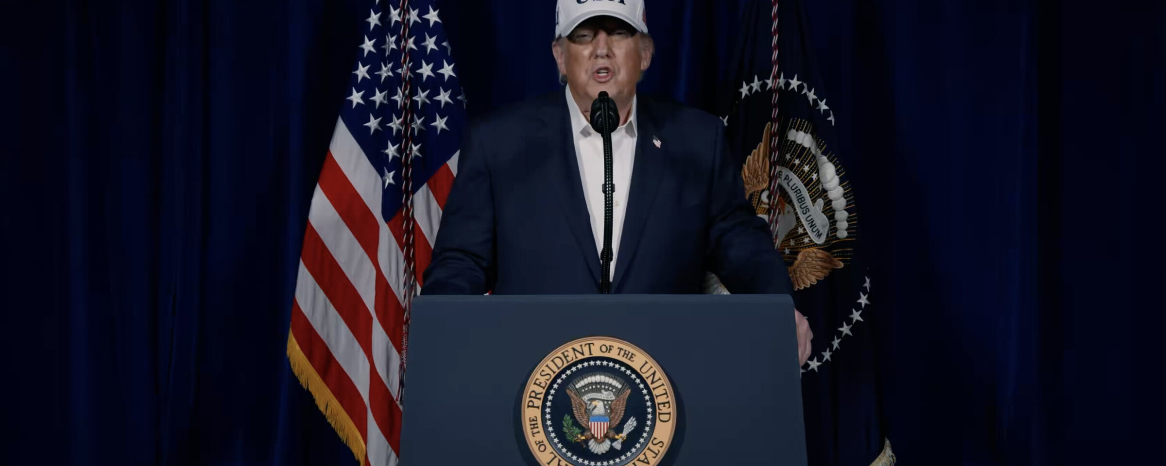 Donald Trump stands at presidential podium speaking and wearing a baseball hat reading "USA"
