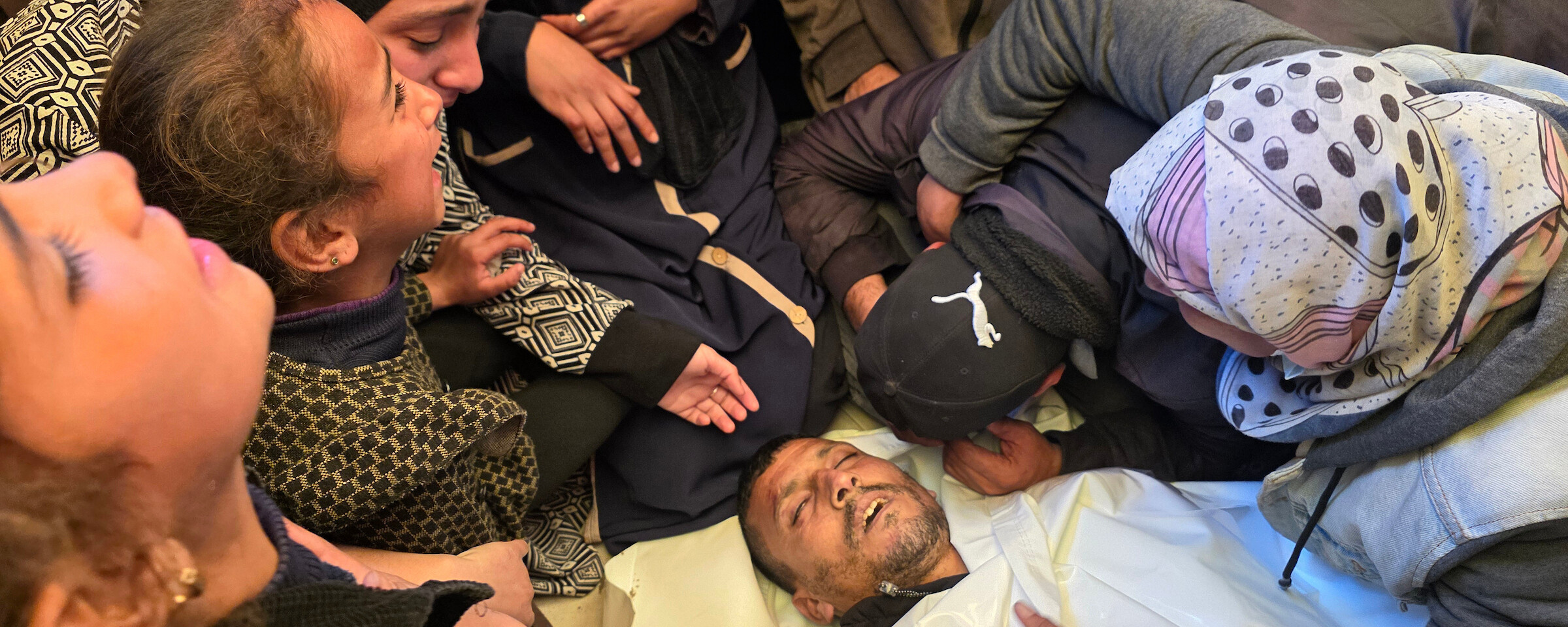 Women and girls mourn over the body of a man wrapped in a white shroud.