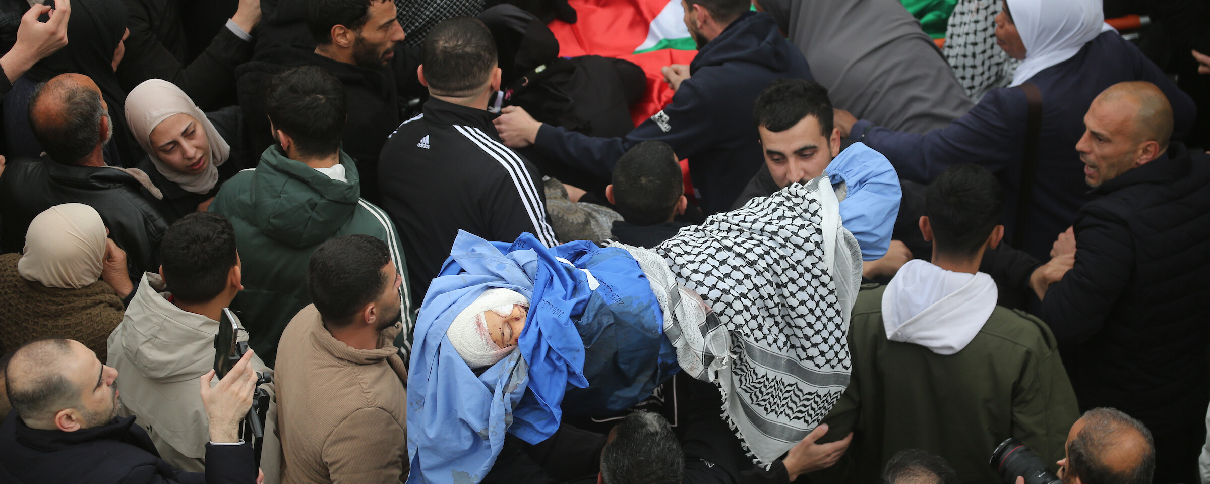 Crowds of mourners gather around the bodies of the Bani Odeh family, wrapped in shrouds, Palestine flags and keffiyehs