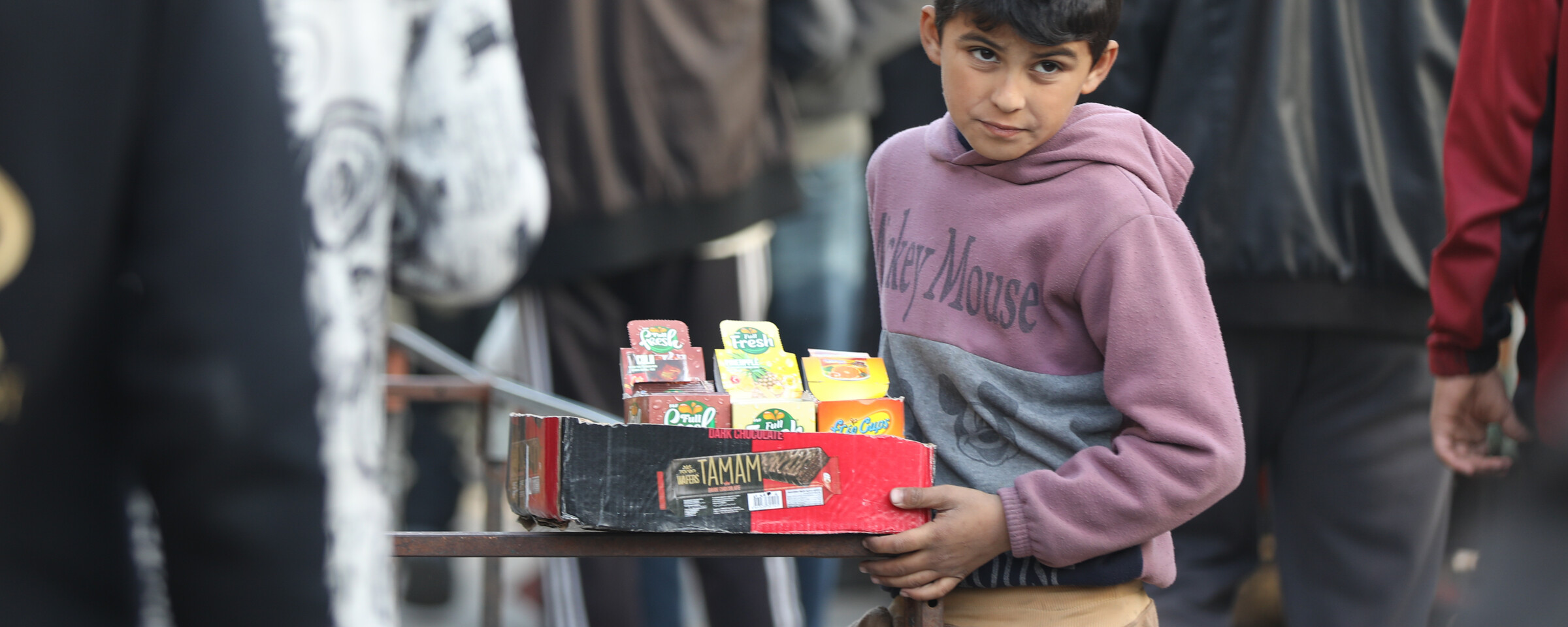 A boy wearing a Mickey Mouse sweatshirt holds a tray of goods.