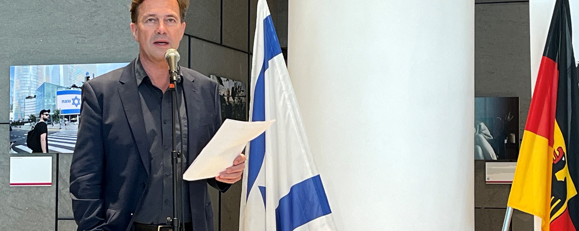 Man holds a piece of paper as he stands behind a microphone stand with flags of Israel and Germany beside him 