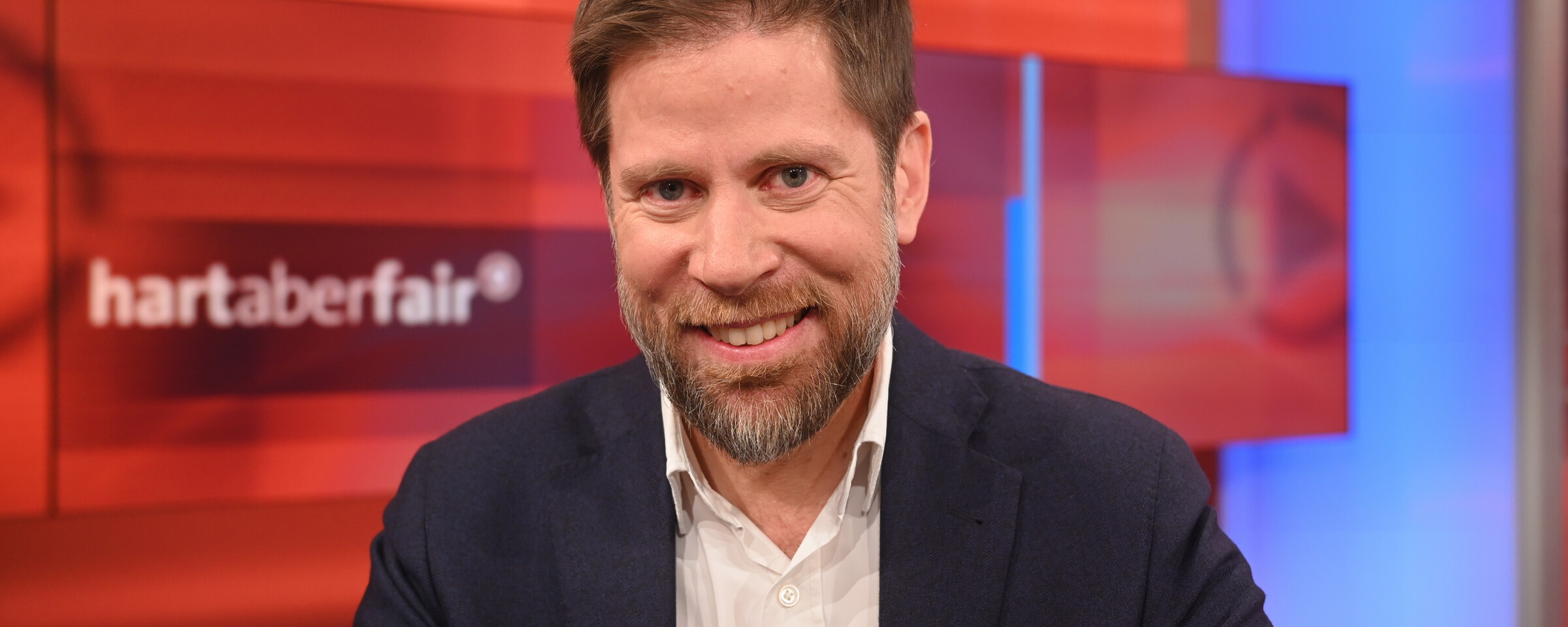 Man wearing a navy jacket and white shirt smiles as he sits in a television studio behind a red backdrop with the words hart aber fair on it. 