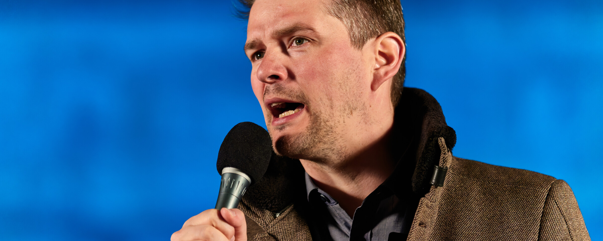 Man speaks into a microphone with a blue backdrop behind him 