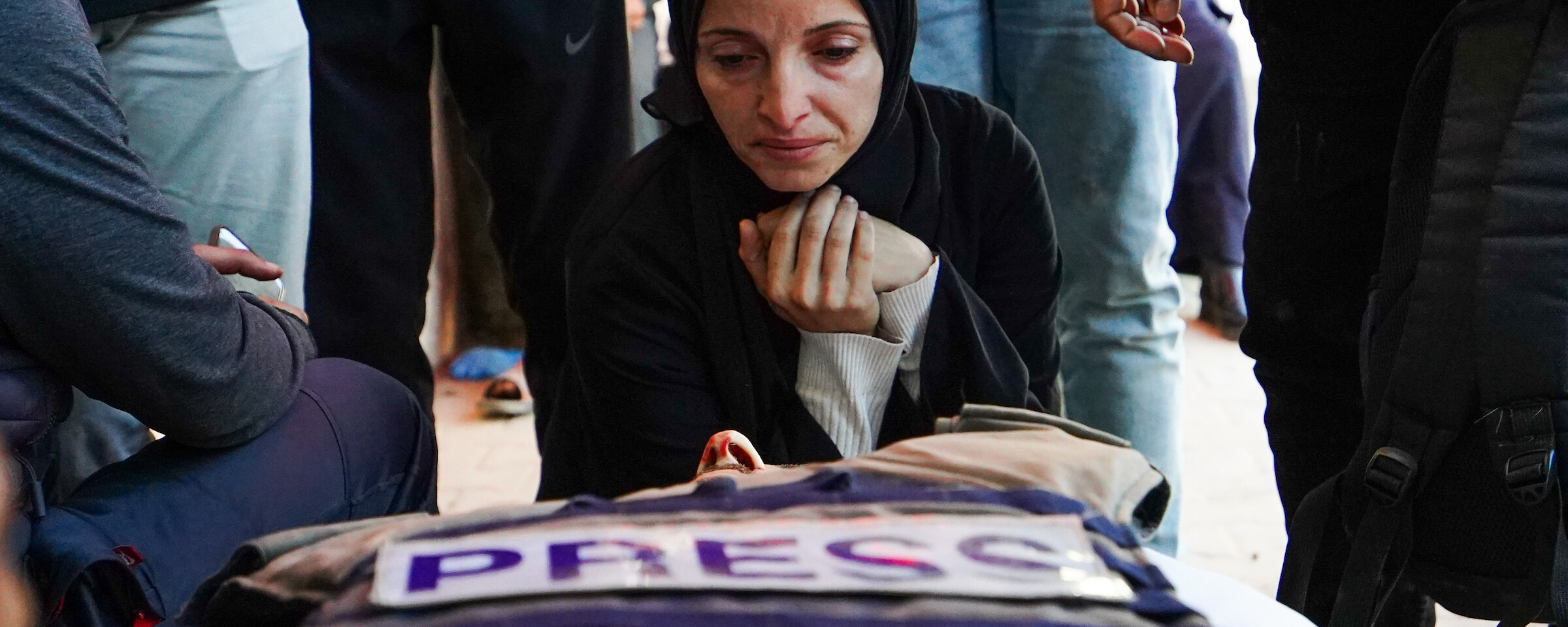 A woman in black clothing kneels beside a body of a man. A press vest is resting on his chest.