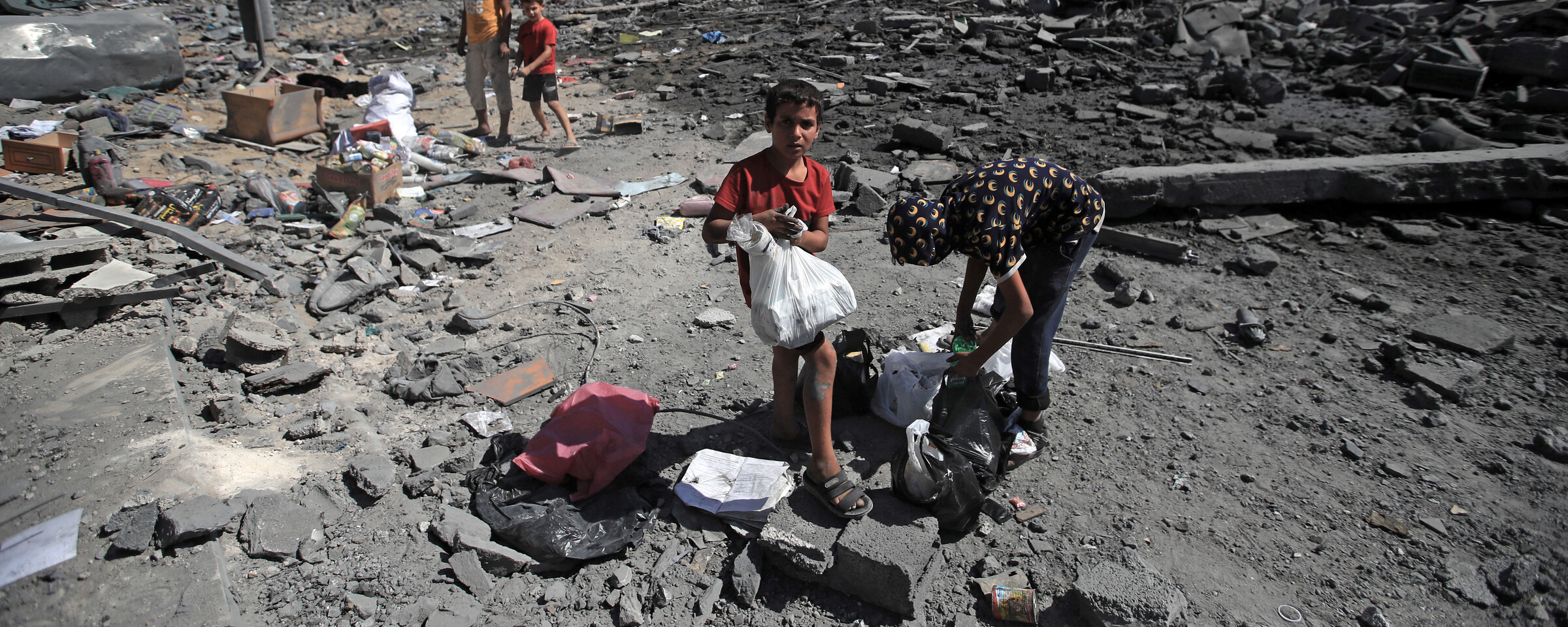A handful of people inspecting through rubble while a boy, holding a plastic bag, is looking at the camera