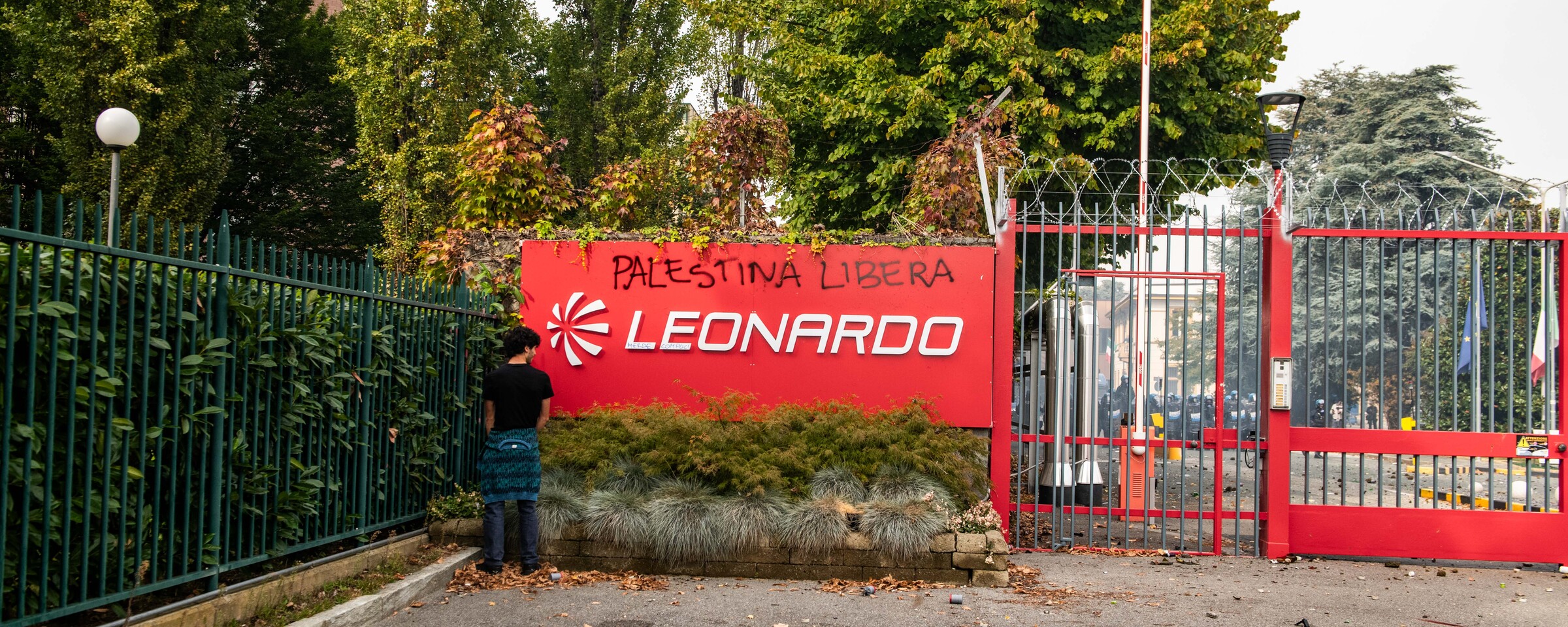 A man stands beside a red sign with the logo of the firm Leonardo and graffiti reading Palestina Libera on it  