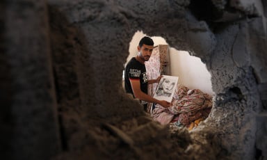 A man holds a drawing through a hole in a wall caused by the Israeli bombing campaign in early August