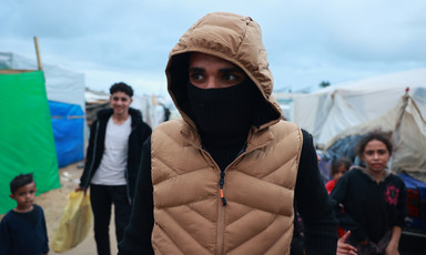 A closeup of a young man wearing a tan puffy hoodie with a mask over his face due to the cold