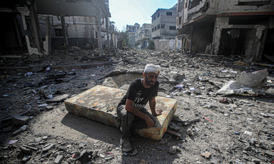 A man with a bandaged head sits on a mattress in a debris-filled street