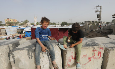 Two boys sit on blocks of concrete with tents behind them 