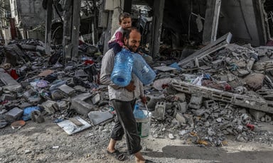 A man carries a child on his shoulders and water containers in his hands as he walks through rubble in southern Gaza 