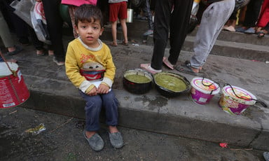 A young Palestinian child sits alongside food as others queue