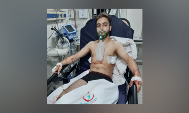 A man sits up in a hospital, with bandaged wounds on his side and a tube in his mouth