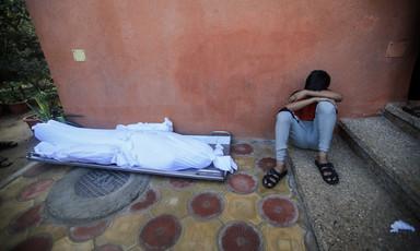 A boy covers his face with his hands while sitting next to three shrouded bodies