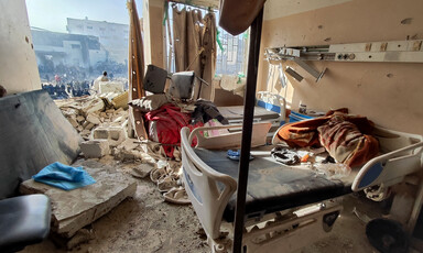 In the daytime, a destroyed hospital room is seen with one of its walls totally missing and with a view of more destruction outside