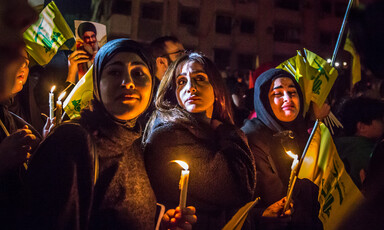 Three young women with sad expressions on their faces hold lit candles and Hizballah flags