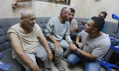 Five Palestinian men seated or squatting with more people in background