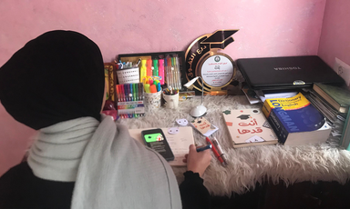 Young woman is seen from back while sitting at desk with a phone, notebook and writing tools