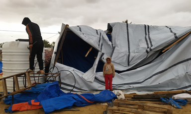 Under cloudy skies, a child stands in front of a collapsed tent