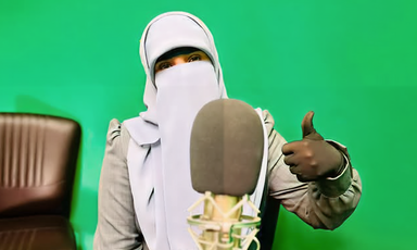 Woman wearing niqab gives a thumbs up while sitting between a microphone and a green screen