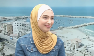 Young woman wearing a jean jacket and mustard yellow hijab smiles slightly while sitting in front of backdrop of Gaza port