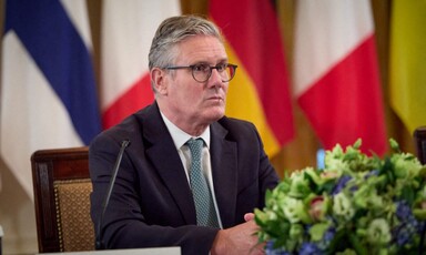 Keir Starmer sits at a table with flowers in front of him and flags behind him