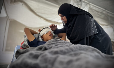A woman leans and holds her hand over a boy on bed with a bandage wrapped around his eyes and head