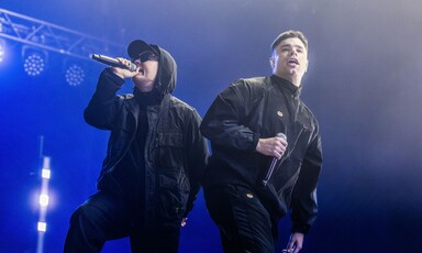 Two members of the group Kneecap wearing black jackets as they perform onstage 