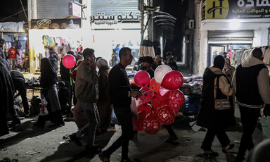 At nighttime, a man holding red balloons walks down a street with shops