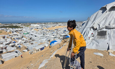 A young boy on crutches stands next to a tent overlooking white tents on sand.
