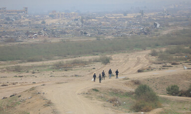 A wide shot of a group of young men walking across a vast expanse of land, with rubble and damaged buildings stretching out before them