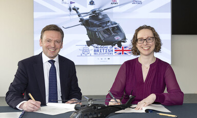 A man and a woman sit on a desk with documents in front of them and with a screen showing an image of a helicopter behind them