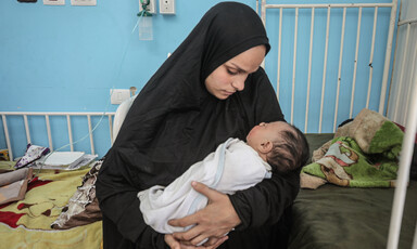 Against a blue wall of a hospital, a woman holds a swaddled infant.