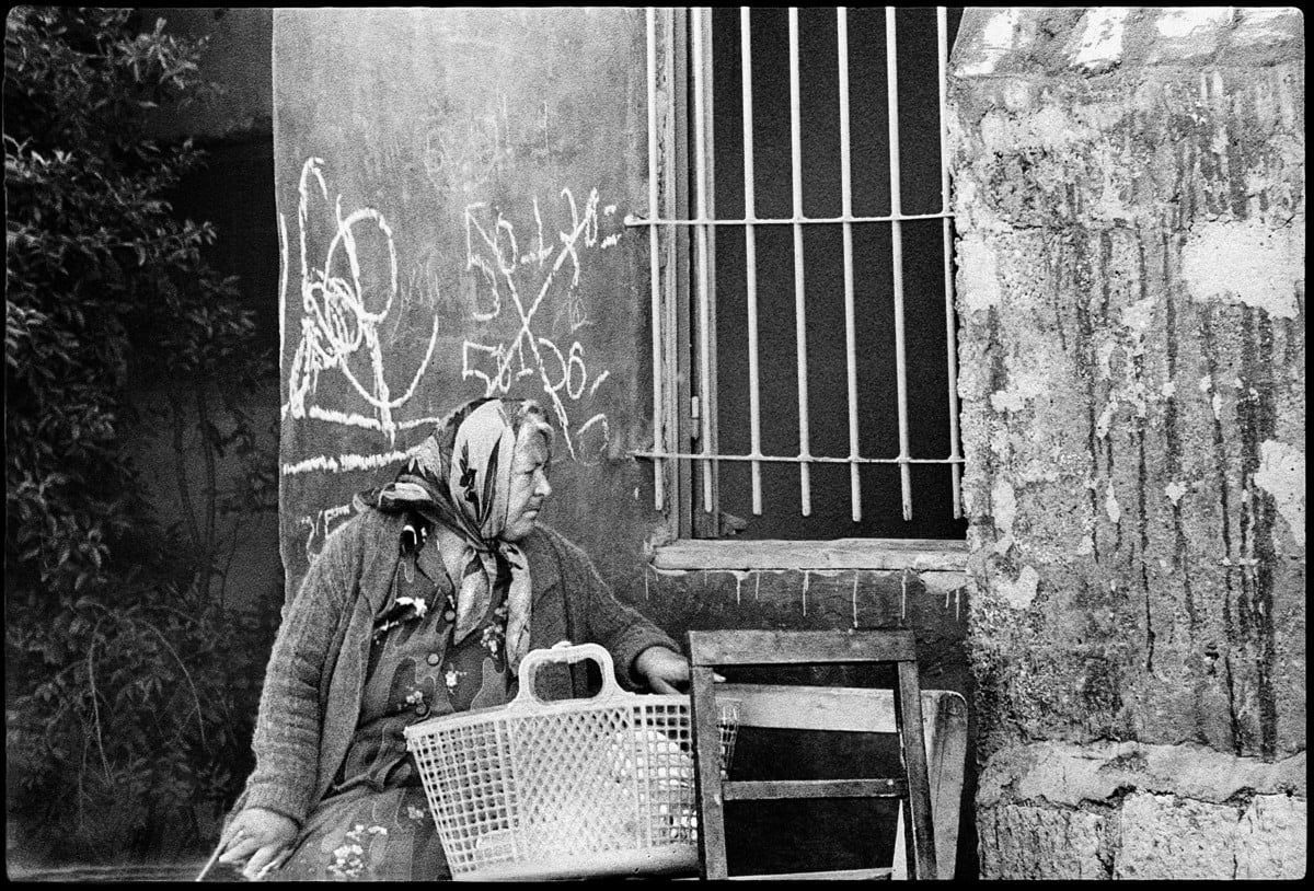 Woman with basket on her lap sits in front of building with a barred window